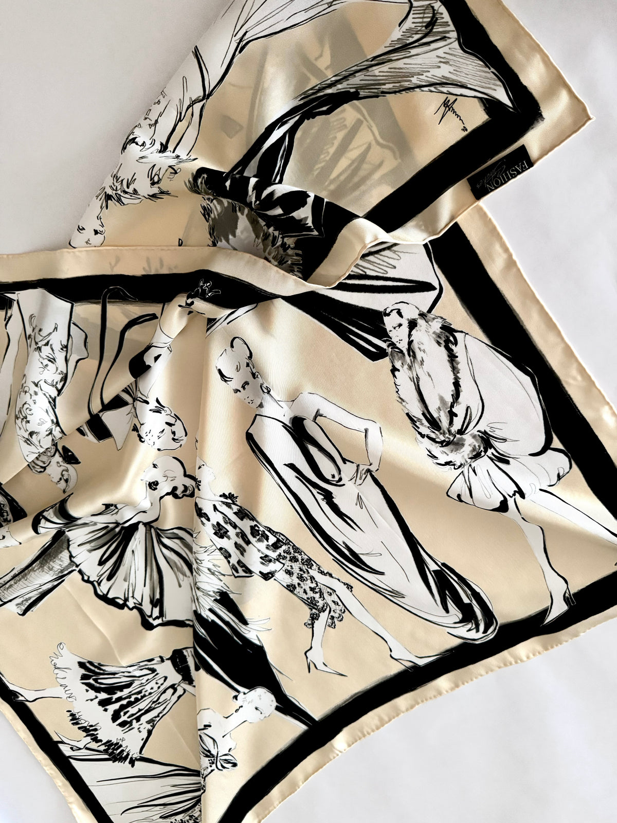 A beige silk scarf with a black and white fashion illustration print, featuring elegant figures and a bold black border, laid out on a white background.